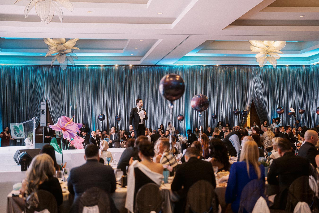 A speaker addressing an audience at a formal event with decorative spherical balloons, elegant lighting, and elaborate drapery, creating a sophisticated atmosphere in a large banquet hall.