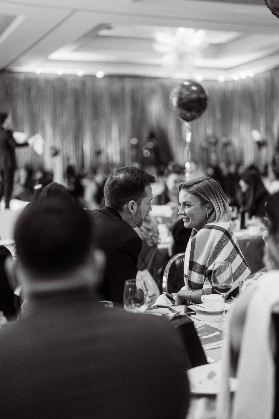 Black and white image of a lively indoor event, with elegantly dressed guests seated at tables, engaging in conversation. The room features soft lighting, decorative balloons, and blurred party decorations in the background, creating a warm and inviting atmosphere.