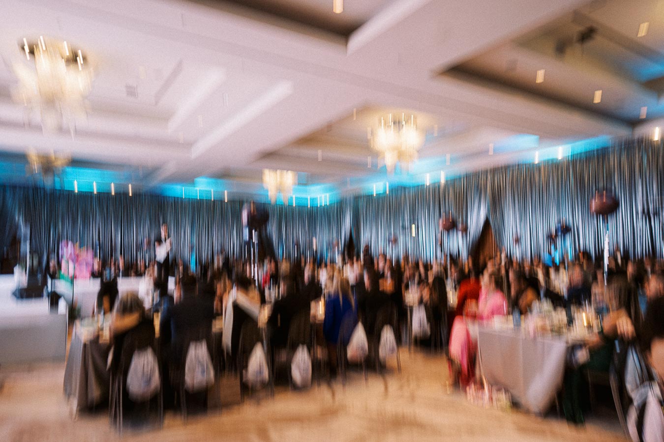 Blurry image of a large indoor event with people sitting at tables, elegant decor, and chandeliers, creating a formal, festive atmosphere.