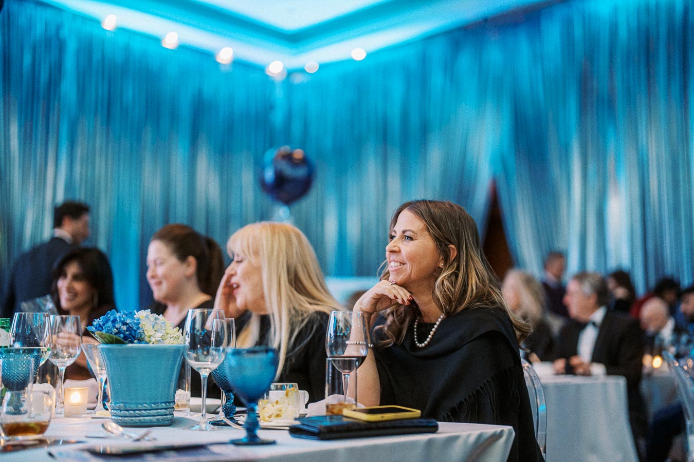 Elegant gala event with guests seated at a table, featuring blue decor and wine glasses, creating a sophisticated atmosphere.