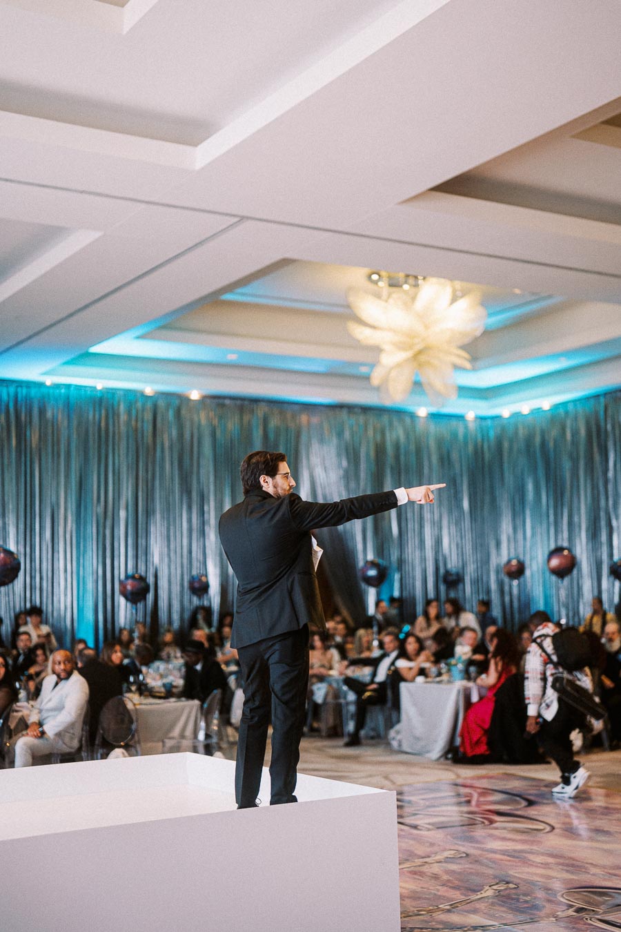 A man in a suit gestures while speaking on a stage at a formal event, with an audience seated at tables under elegant lighting and decor.