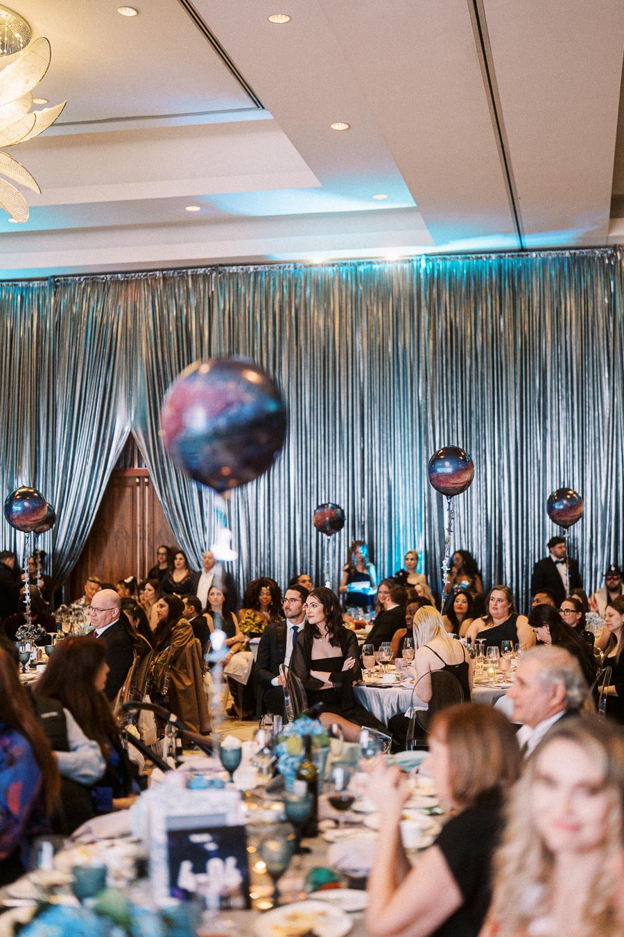 A formal event with attendees seated at elegantly decorated tables, featuring a shimmering backdrop and unique balloon centerpieces.