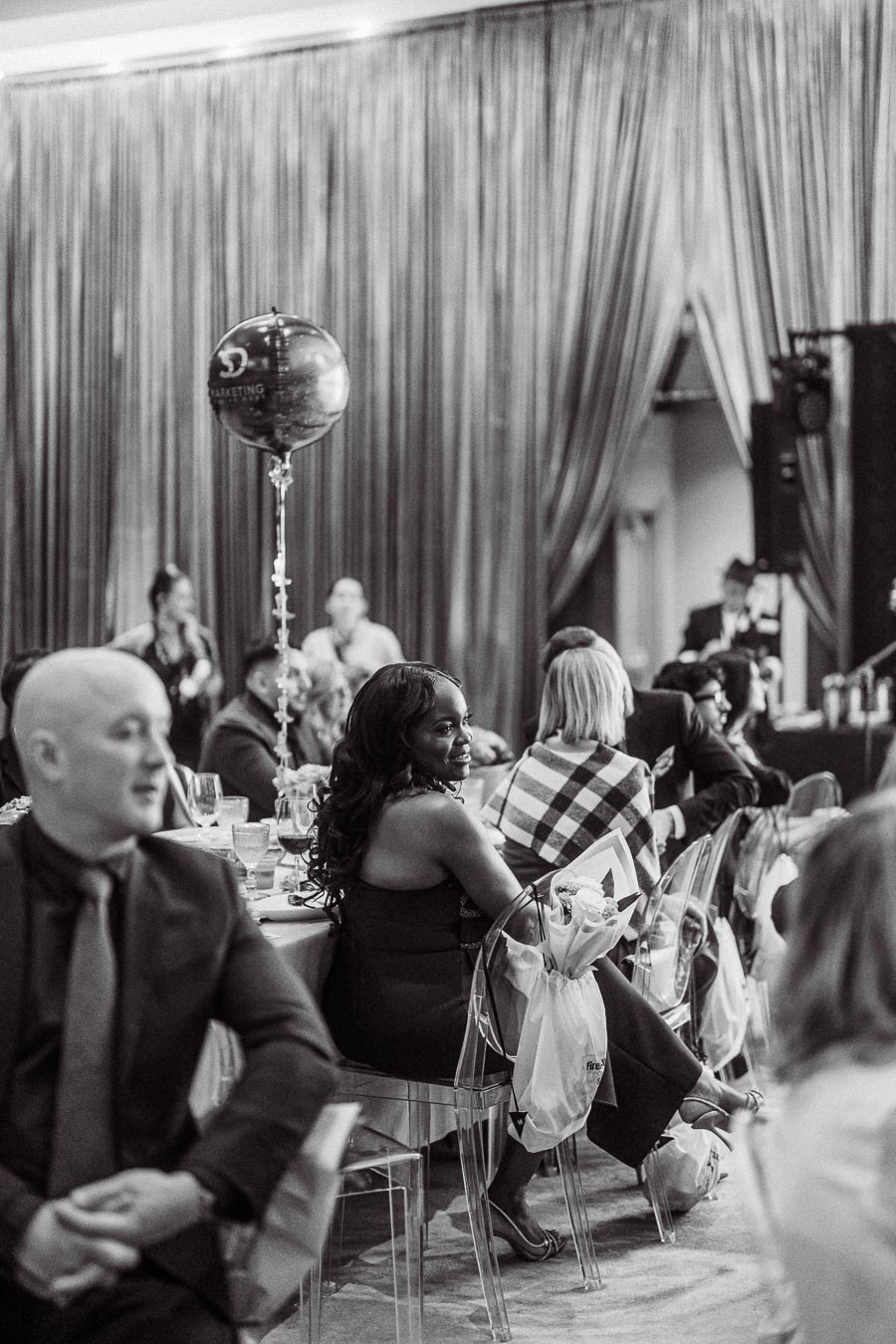 Black and white image of an elegant indoor event, featuring people seated at tables and engaging in conversation. A balloon floats above one table, adding a festive touch. The atmosphere is lively, with guests dressed in formal attire under the soft glow of drapery.