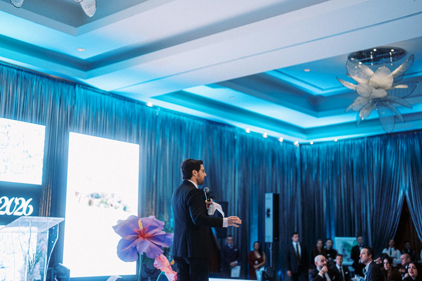 Speaker presenting at a conference in a blue-lit room with audience attendees in formal attire.