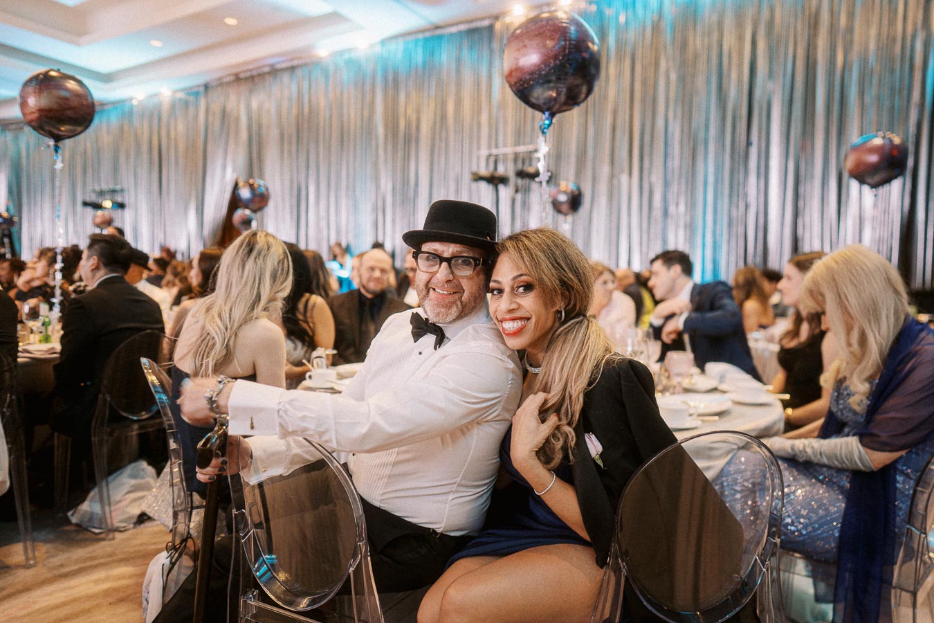 A joyful couple at an elegant gala event, seated and smiling at a table adorned with decorative balloons, surrounded by an elegantly dressed crowd in a softly lit, glamorous venue.
