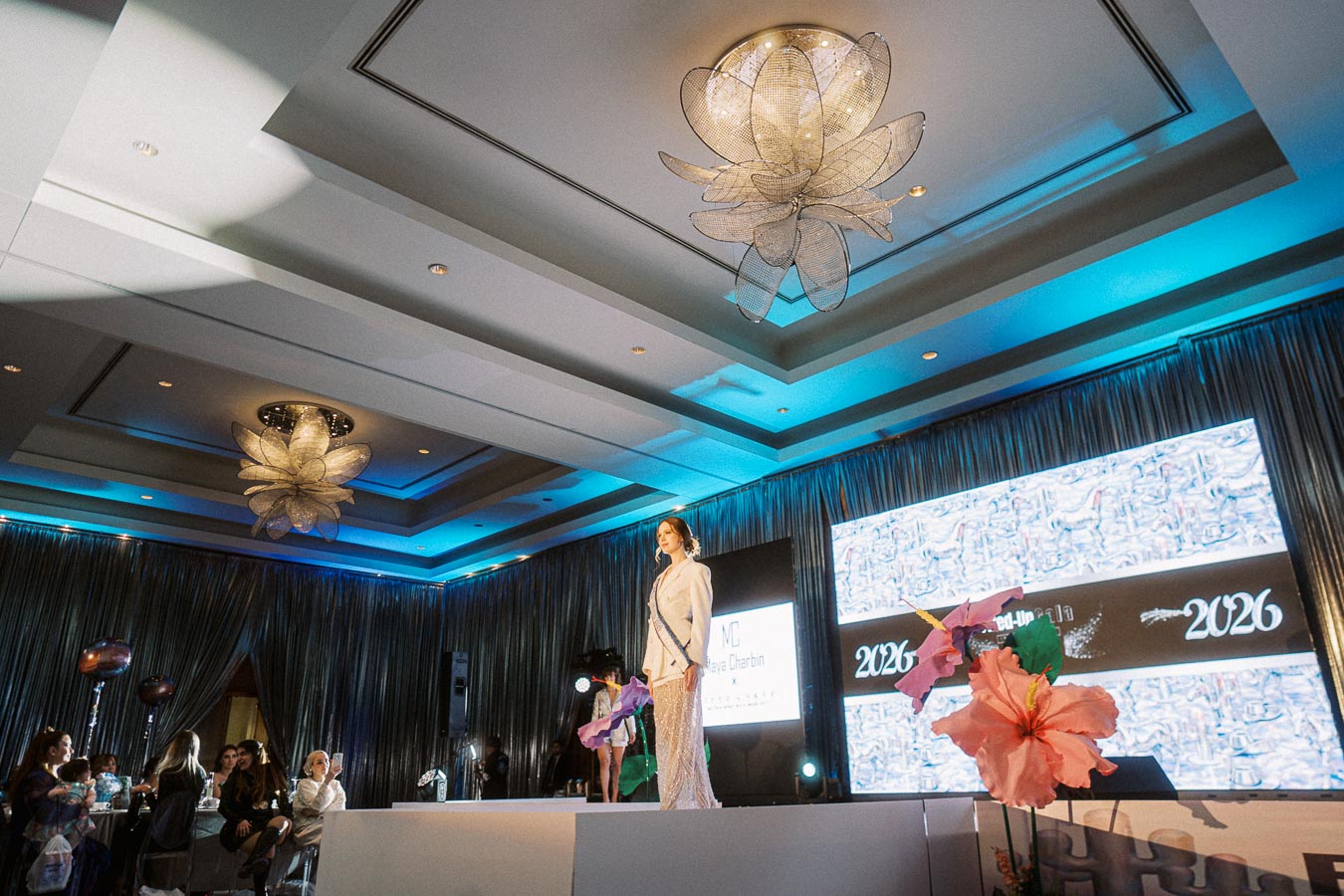 Fashion runway event inside a modern venue with elegant lighting, featuring a model in a stylish outfit on stage, surrounded by attendees.