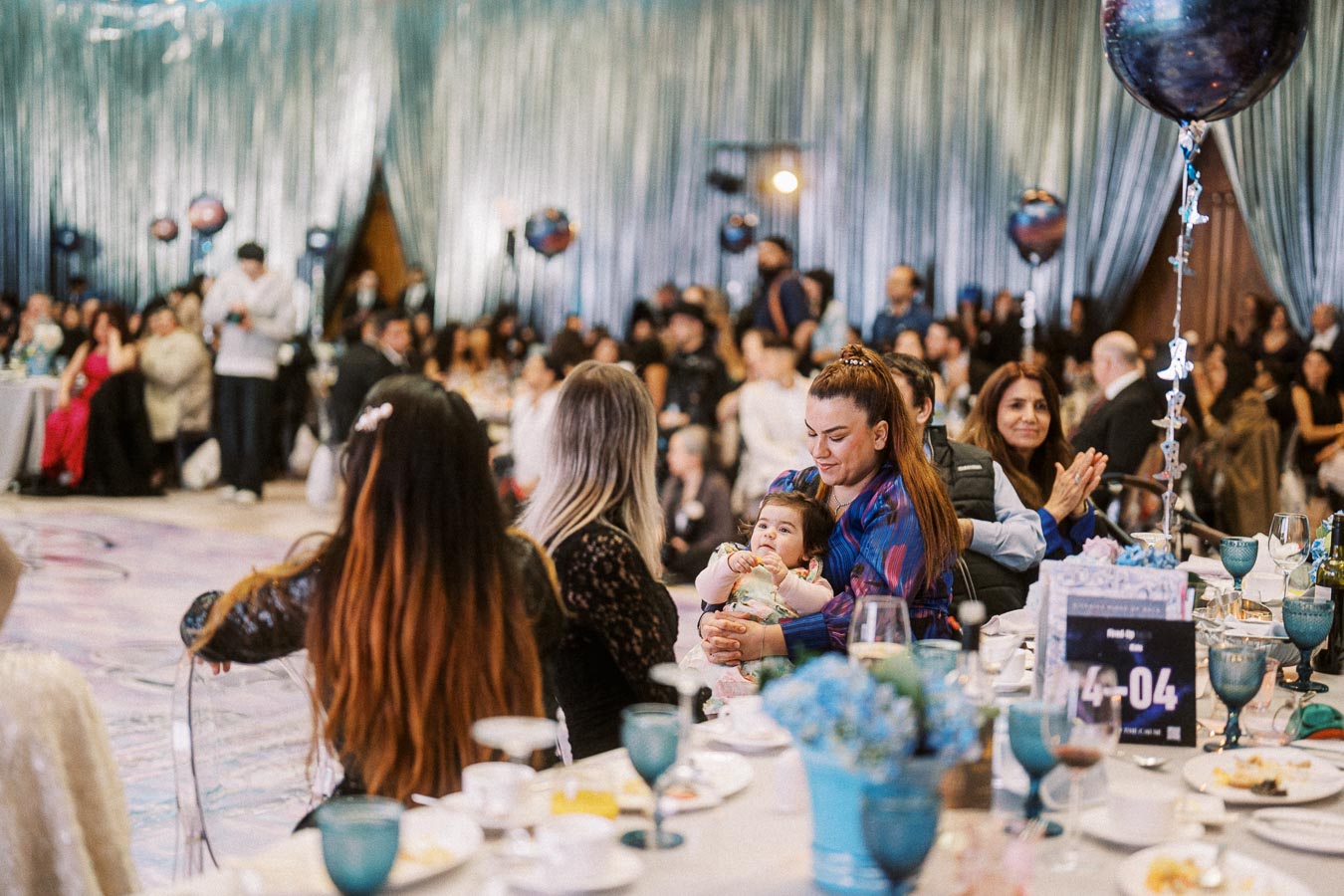 A lively celebration in a decorated banquet hall, with guests seated at elegantly set tables. A smiling woman holds a baby, surrounded by attendees engaged in conversation and clapping, with festive balloons and curtains in the background.