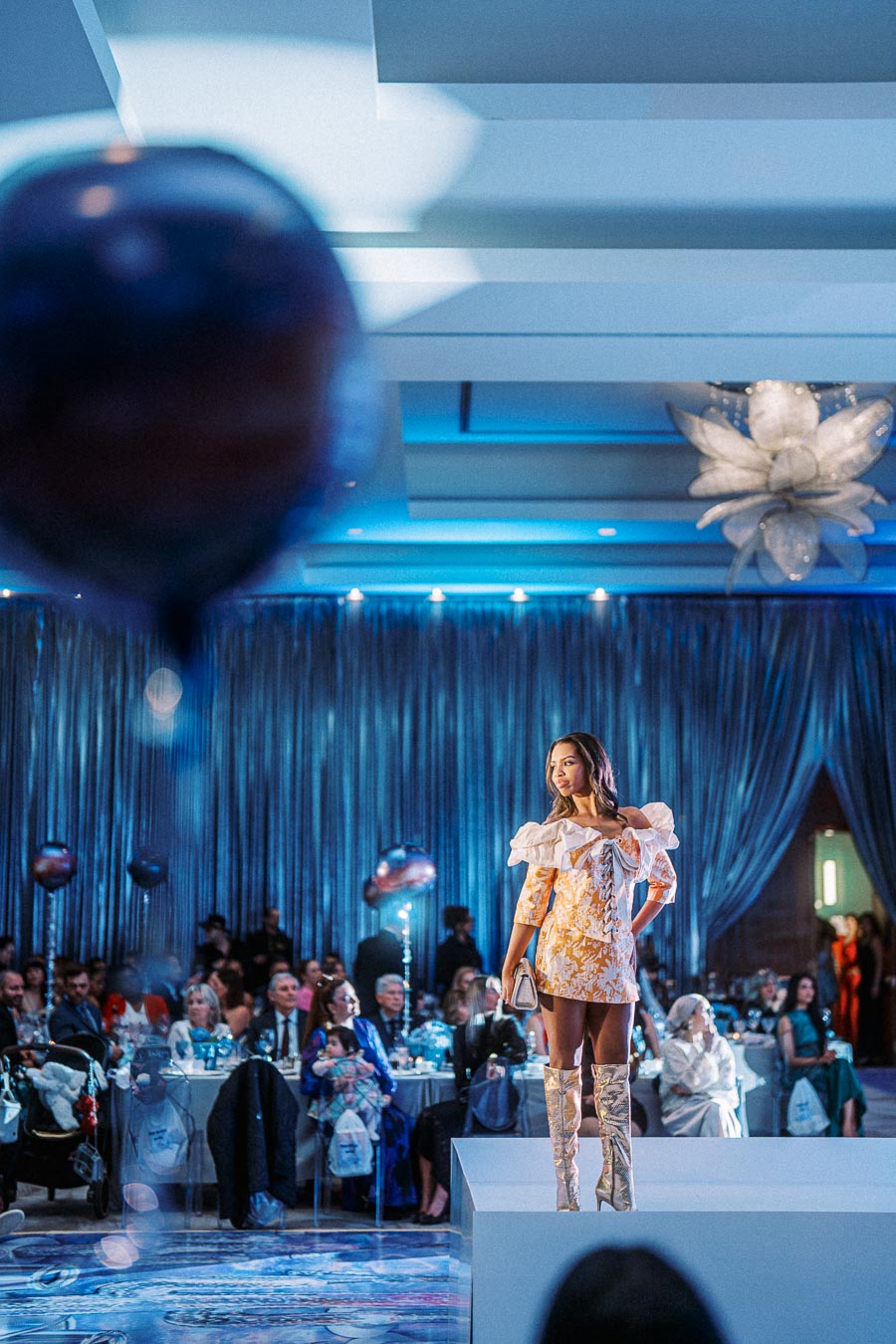 Model walking the runway in a fashionable orange and white dress with ruffle details and knee-high boots at a glamorous indoor fashion event, surrounded by an audience and stylish decor.
