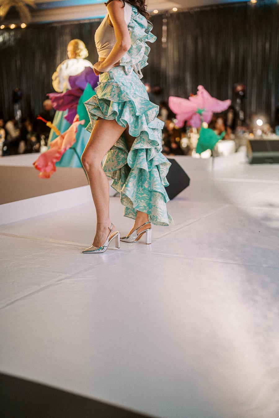 Fashion model wearing a turquoise ruffled high-low dress and silver heels on a runway.