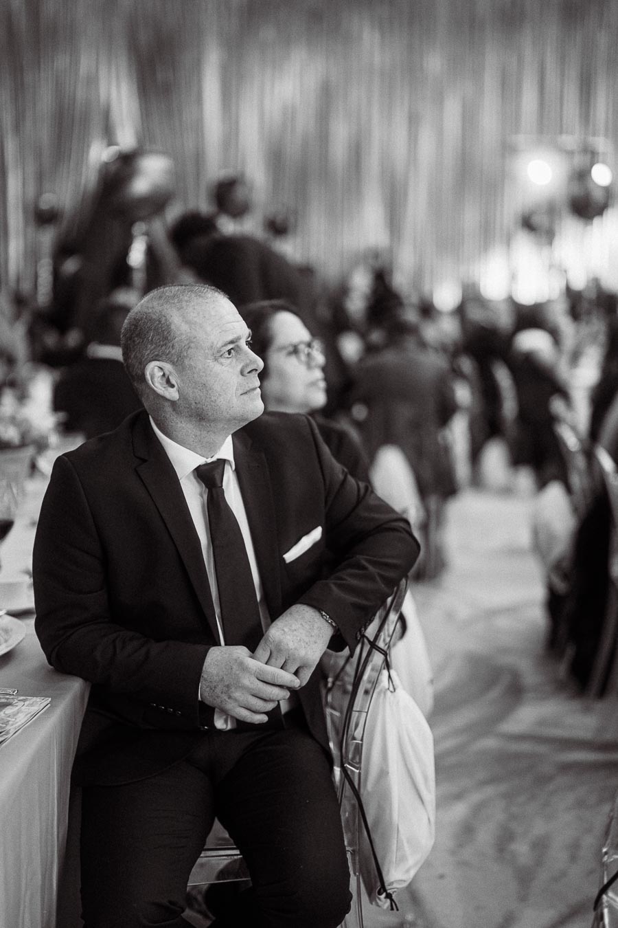 Black and white photo of a man in a suit attentively sitting at a formal event, with blurred background showcasing a crowded gathering.