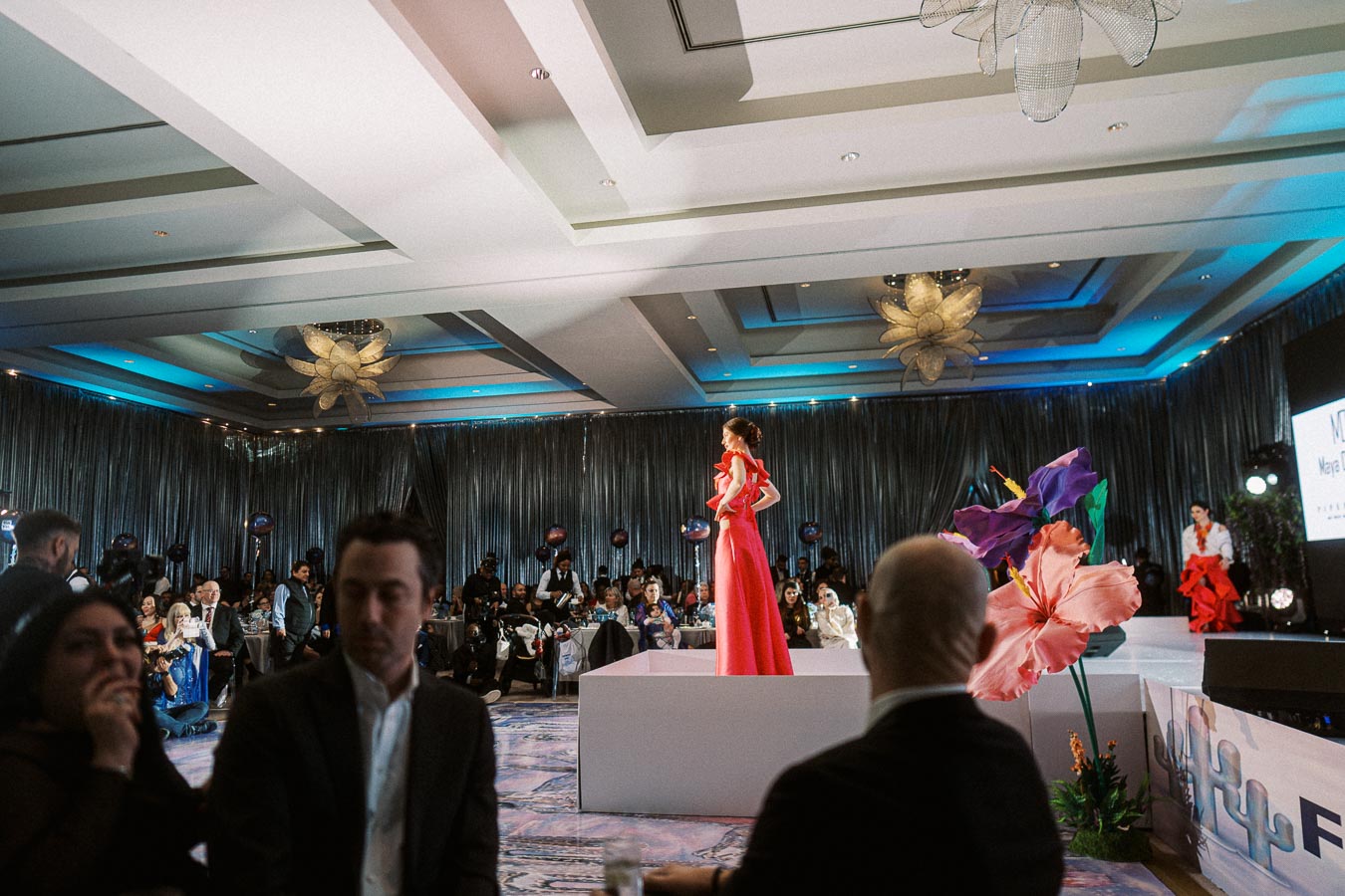 Fashion show event with a model in a red dress walking on a runway, surrounded by an audience seated in an elegantly decorated venue, featuring large, colorful floral decorations.