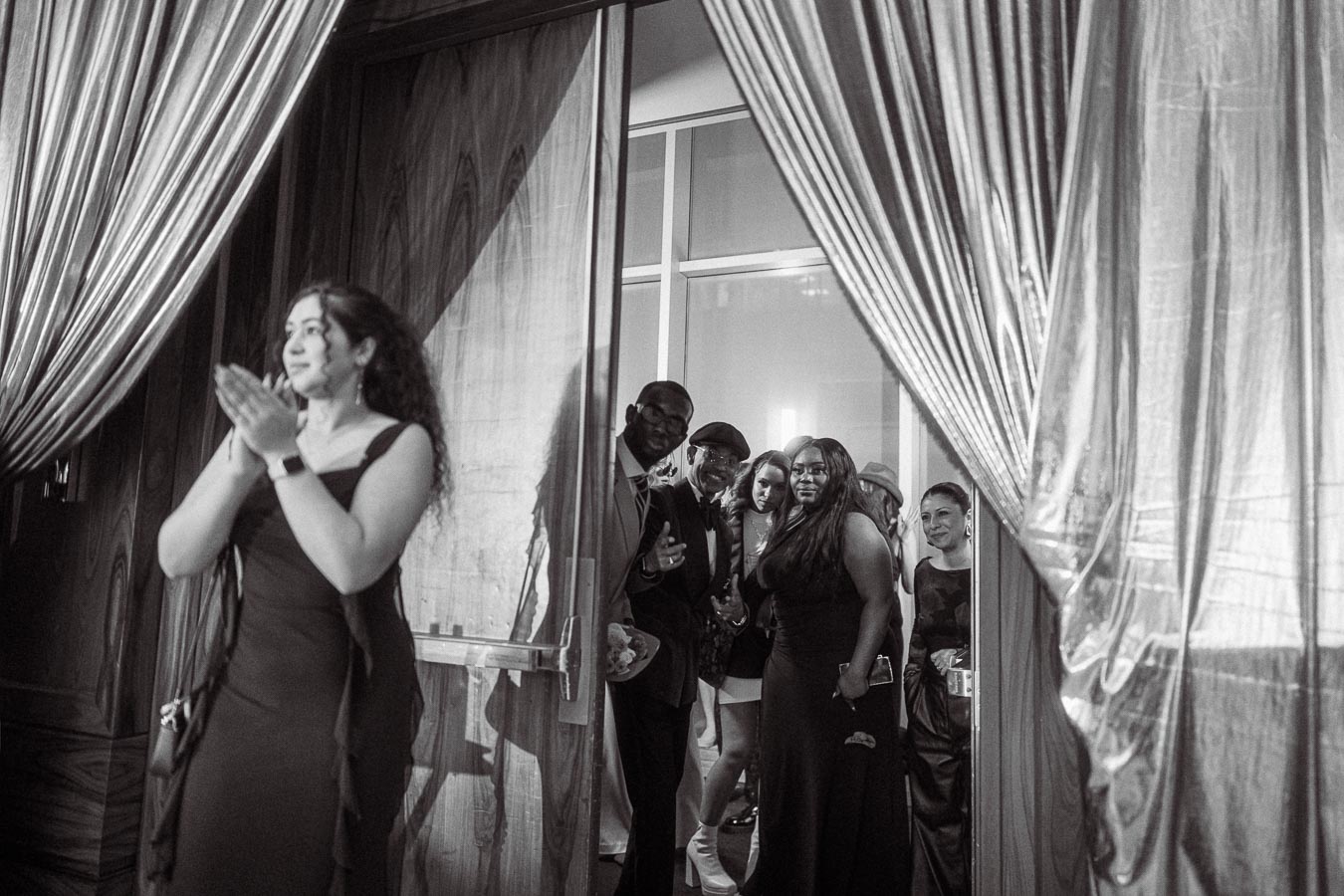 A group of elegantly dressed people gathered at an event entrance, with a woman in a formal gown applauding in the foreground. The setting features luxurious curtains and a stylish atmosphere, captured in black and white photography.