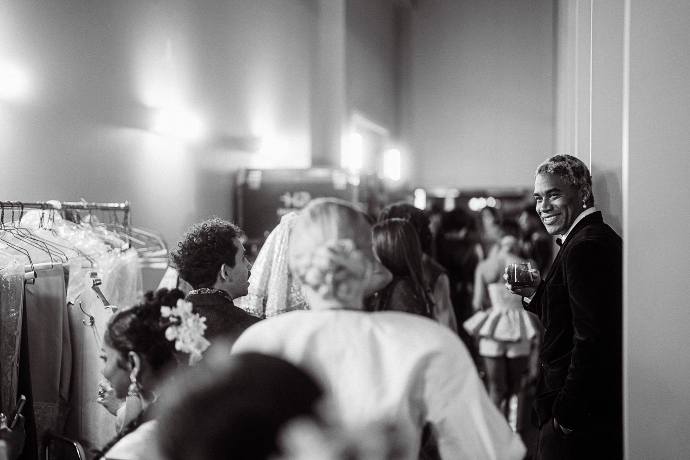 Black and white photo of a stylishly dressed person smiling and holding a drink, standing against a wall in a bustling, crowded room full of people, some examining clothing on a rack.