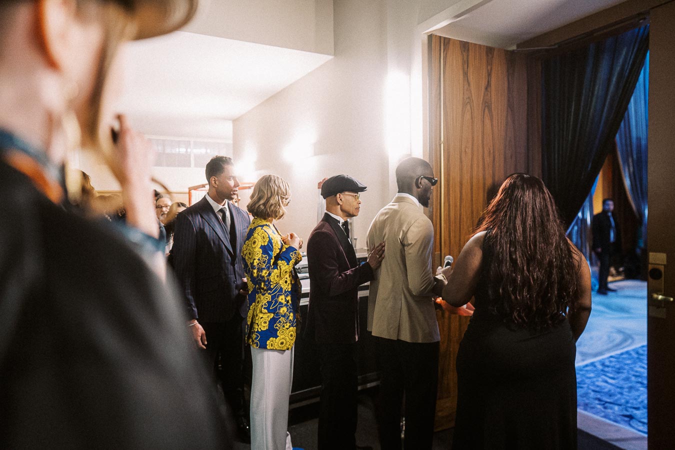 A group of people, dressed in formal attire, stand in line backstage at a professional event, preparing to enter a room through a wooden door, with a carpeted floor visible beyond.