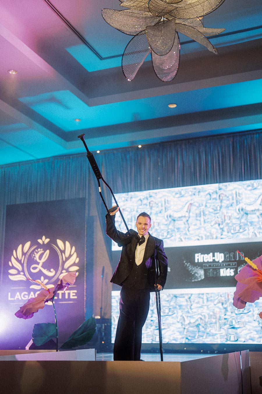 Man in formal attire holding a crutch on stage at an elegant event with vibrant lighting and decorative elements.