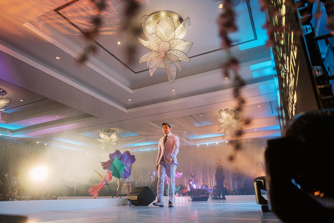 Young man in a stylish light purple suit walks confidently on a modern, brightly lit stage adorned with floral decorations, set against a backdrop of elegant ceiling designs and colorful spotlights at a fashion event.