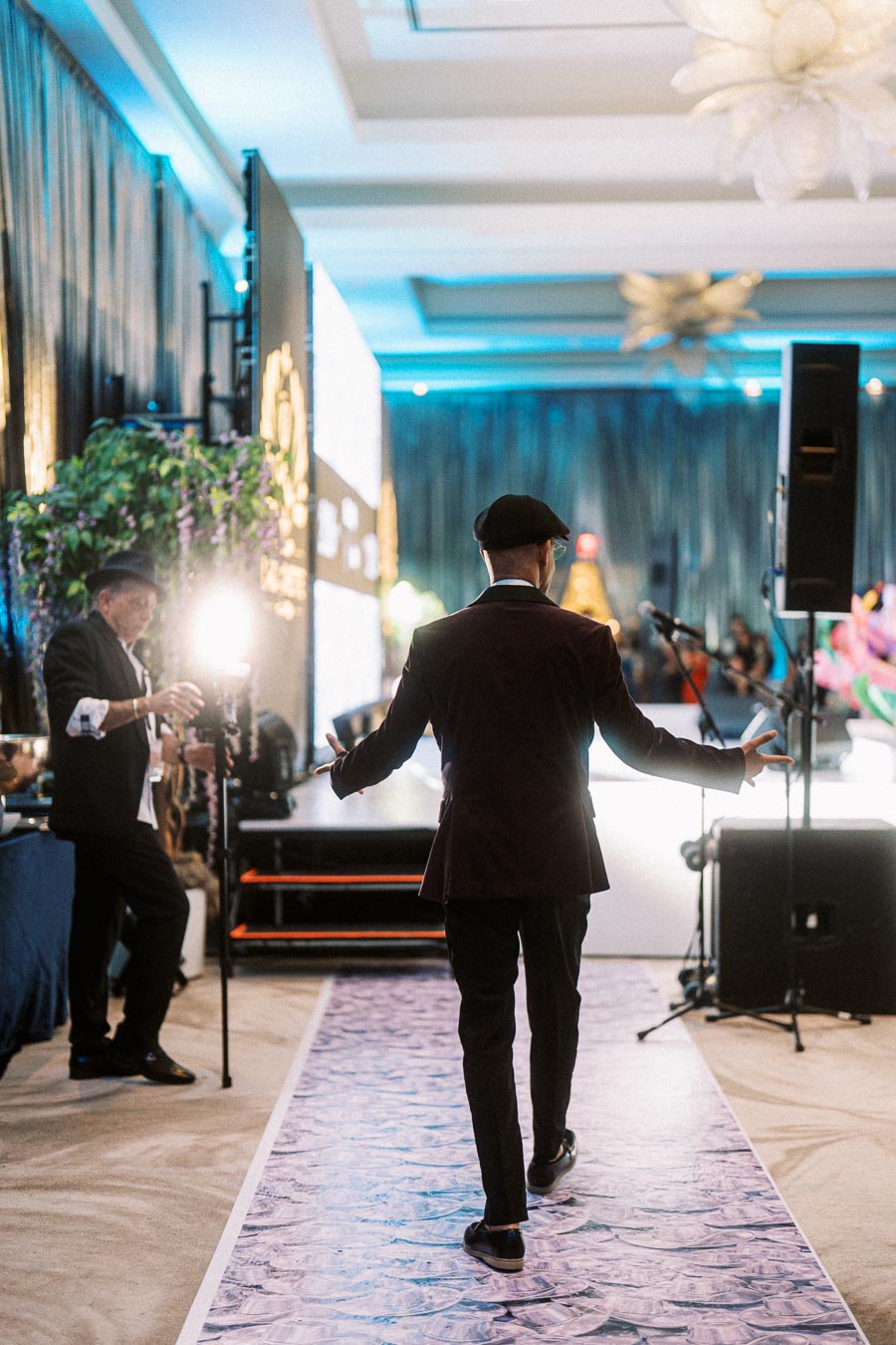 A man in a stylish suit and hat walking down an elegantly decorated event runway with a photographer capturing the moment and vibrant lighting in the background.