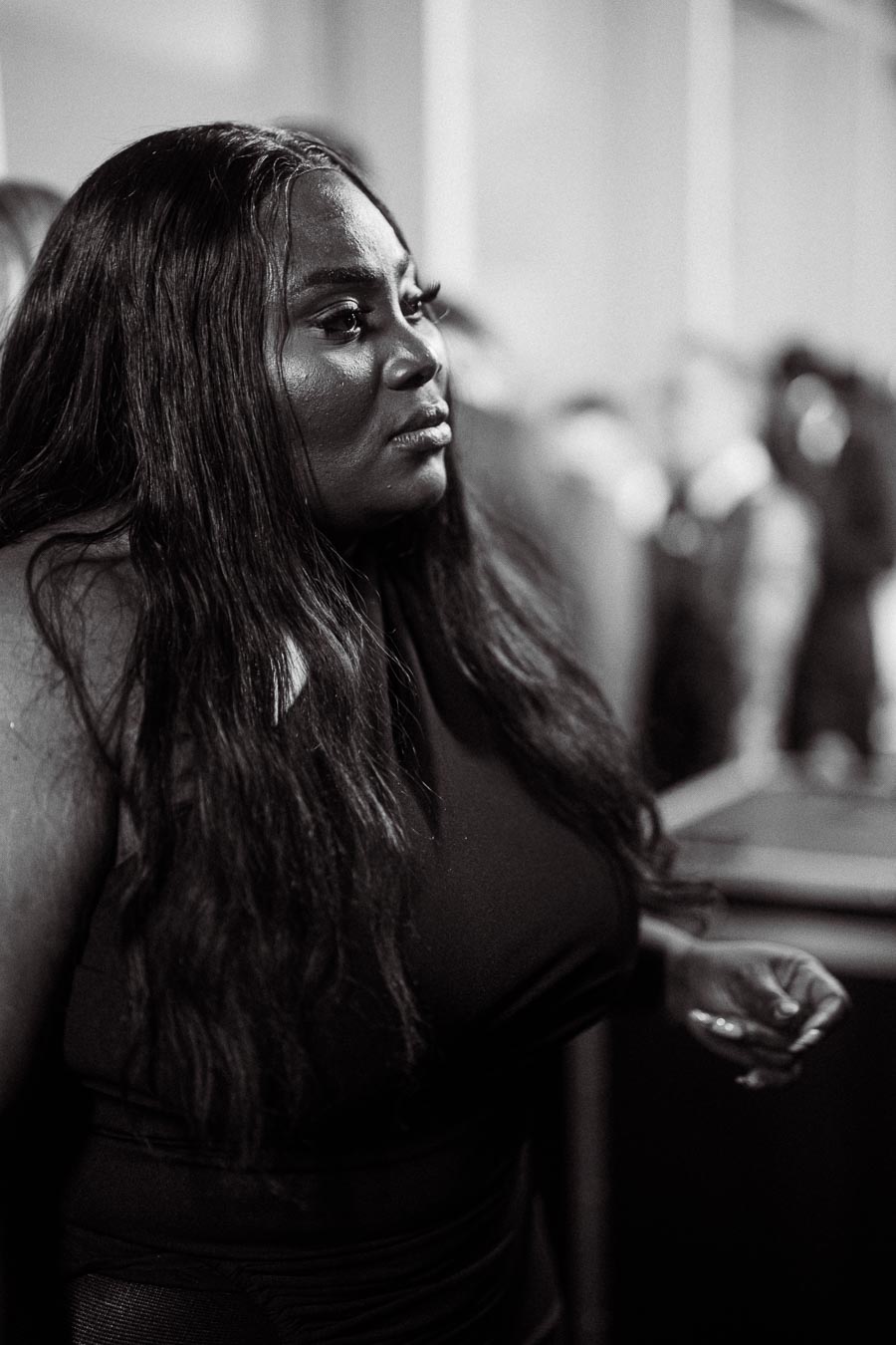 Black and white photo of a woman with long hair in a reflective moment at an indoor event. The blurred background hints at a busy atmosphere.