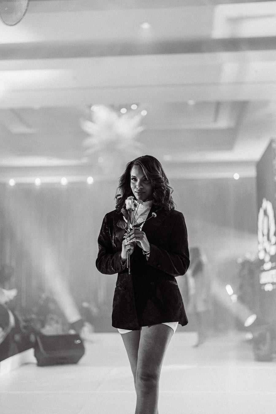 Black and white photo of a model walking on a runway, holding a rose, wearing a stylish blazer at a fashion event.