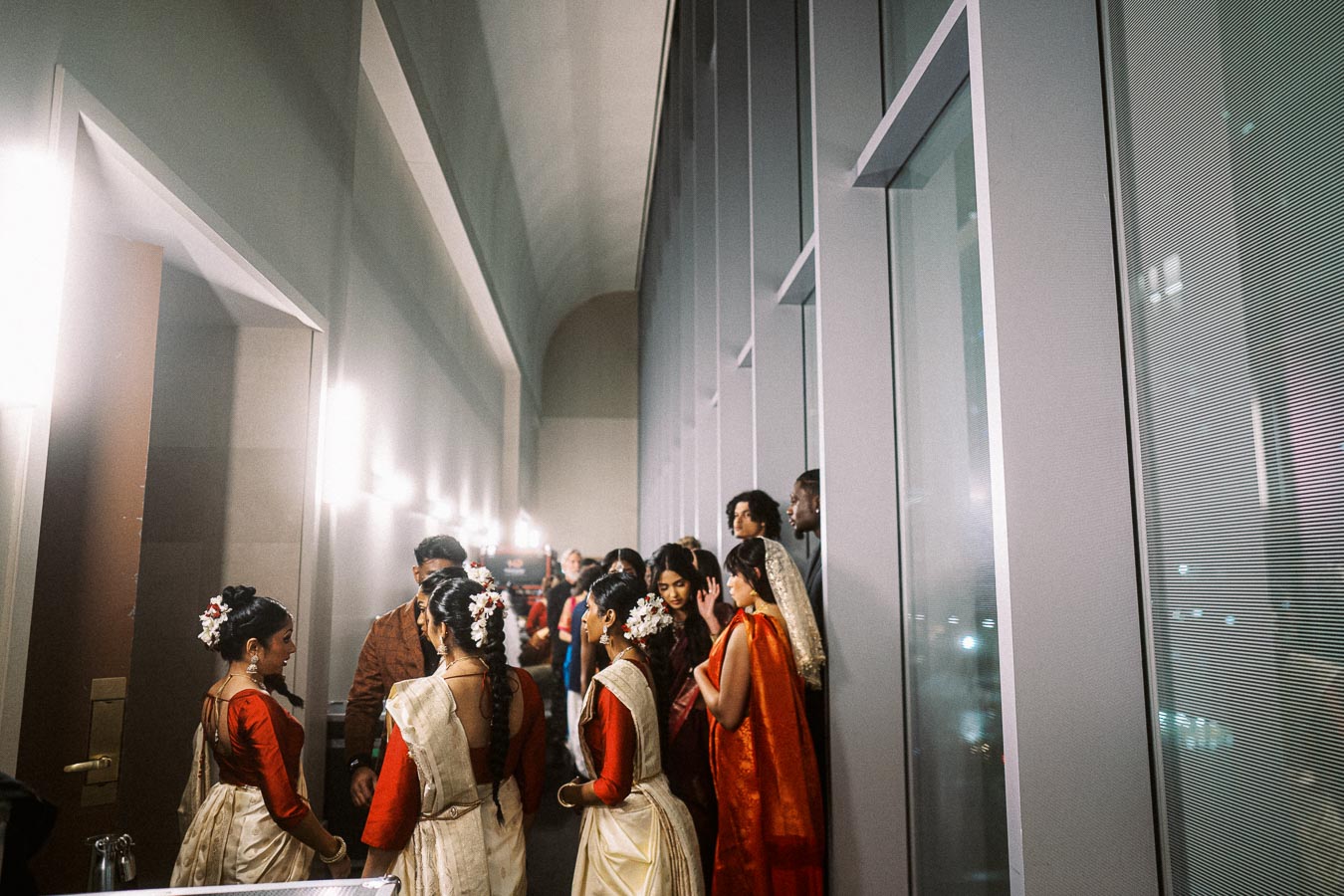 People in traditional attire gather in a modern hallway, preparing for an event. The women wear red blouses with cream sarees adorned with flowers, creating a vibrant cultural scene. The setting features sleek architectural elements, blending tradition with contemporary design.