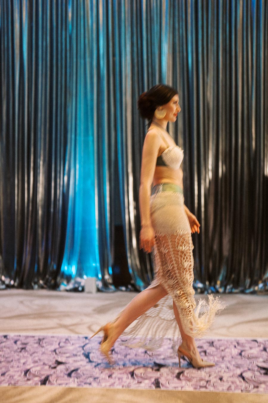 A model in a stylish crochet outfit walks down the runway with a metallic curtain backdrop, highlighting fashion trends at a contemporary fashion show.