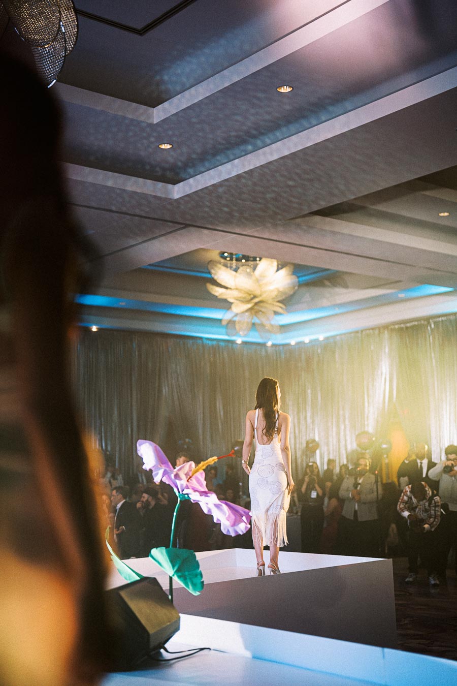 A model in an elegant white dress walks down a fashion runway adorned with large, colorful floral decorations, with a captivated audience and photographers capturing the moment.