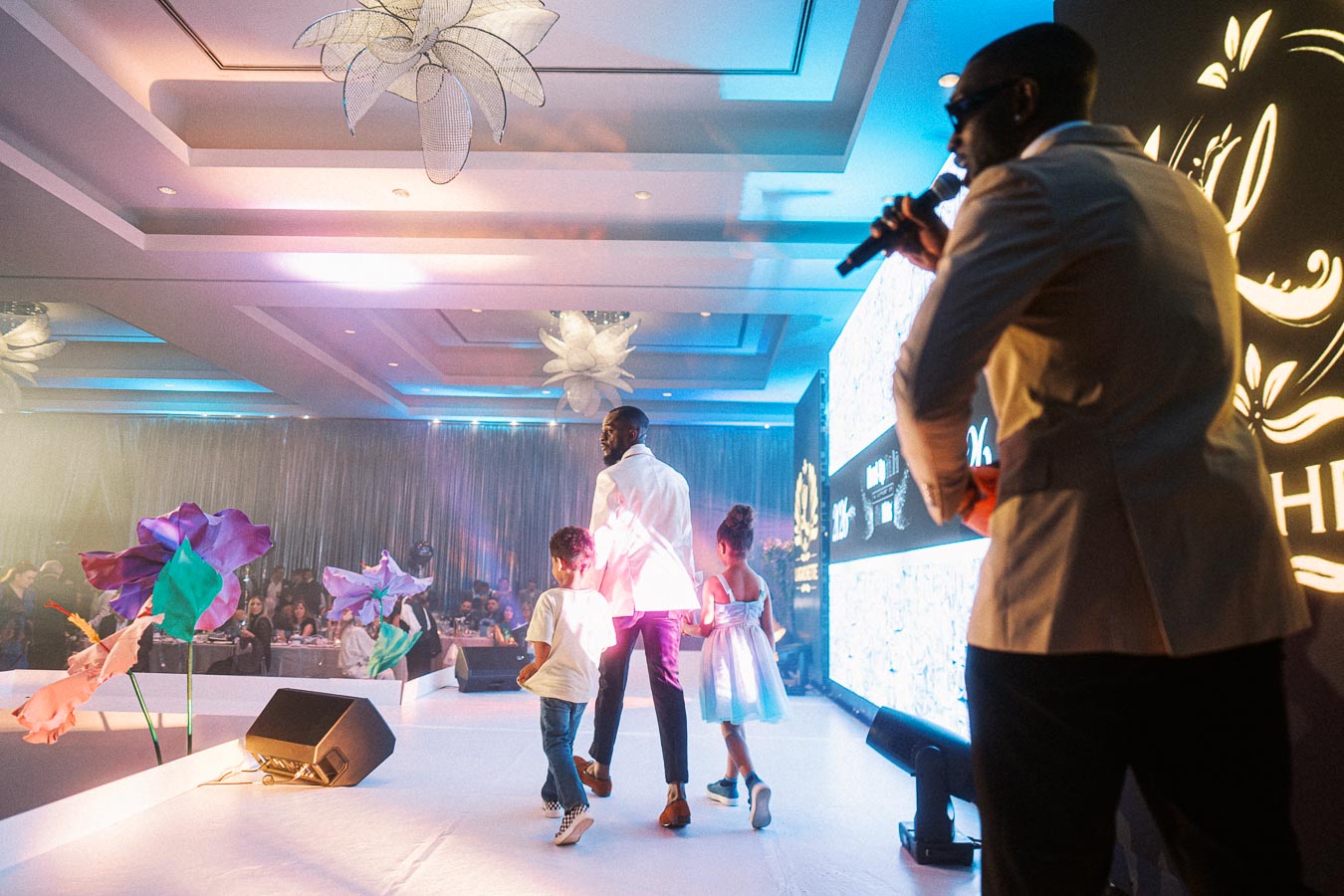 Multicultural fashion event with a stylish host and children walking on stage, surrounded by vibrant floral decor and a modern audience.