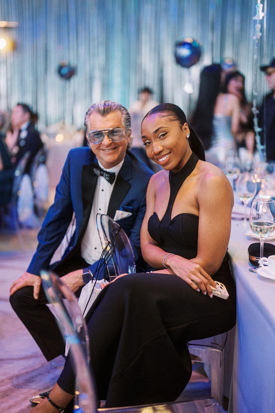 Two elegantly dressed individuals smiling and posing together at a formal event with a festive, colorful setting in the background.