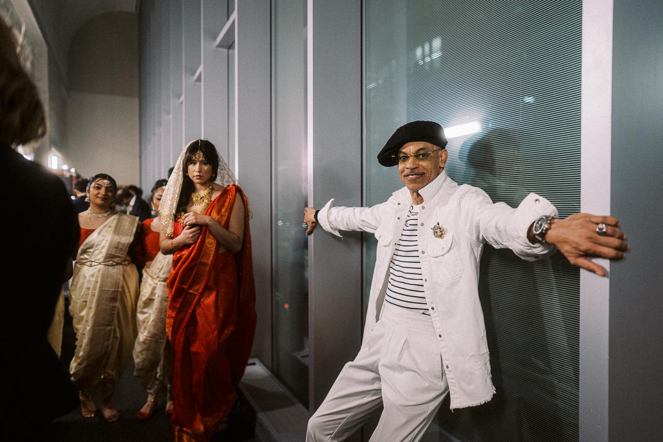 A group of people dressed in vibrant traditional and contemporary fashion at an indoor event, featuring a person in a bright red sari with gold details and another person in a stylish white outfit with a black beret.