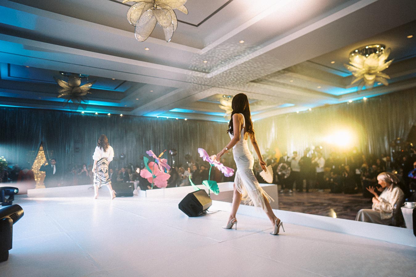 Fashion show runway with models walking in elegant dresses, surrounded by an audience in a stylishly lit venue.