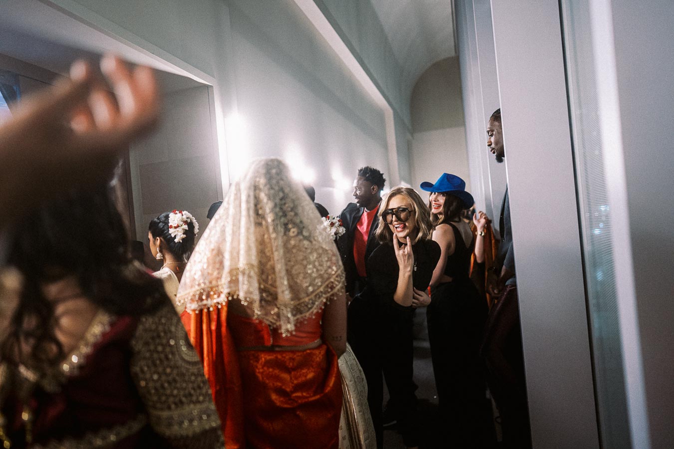 A diverse group of people celebrating indoors, with a focus on a woman wearing sunglasses making a hand sign, while another person dressed in cultural attire with a veil is visible in the foreground. Bright lighting and candid joyful expressions create a festive atmosphere.