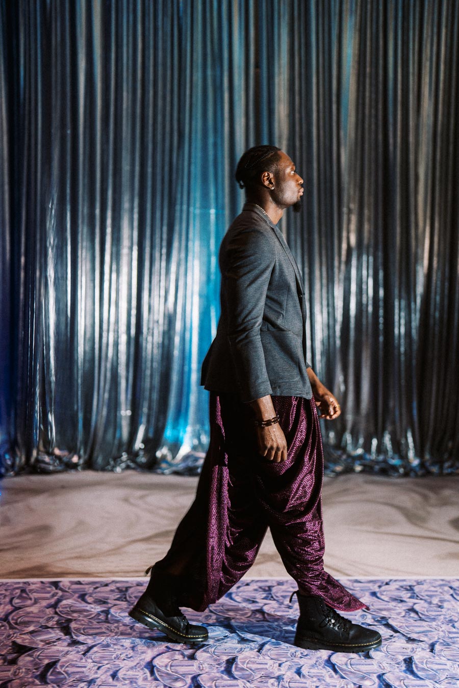Man walking on stylish purple carpet in a modern fashion outfit with a sleek grey blazer and unique purple pants, set against a metallic silver curtain background.