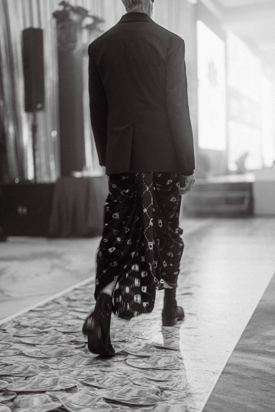Black and white photo of a fashion model walking on a runway covered in dollar bills, wearing a stylish suit jacket and patterned pants, creating a luxurious and artistic fashion show atmosphere.