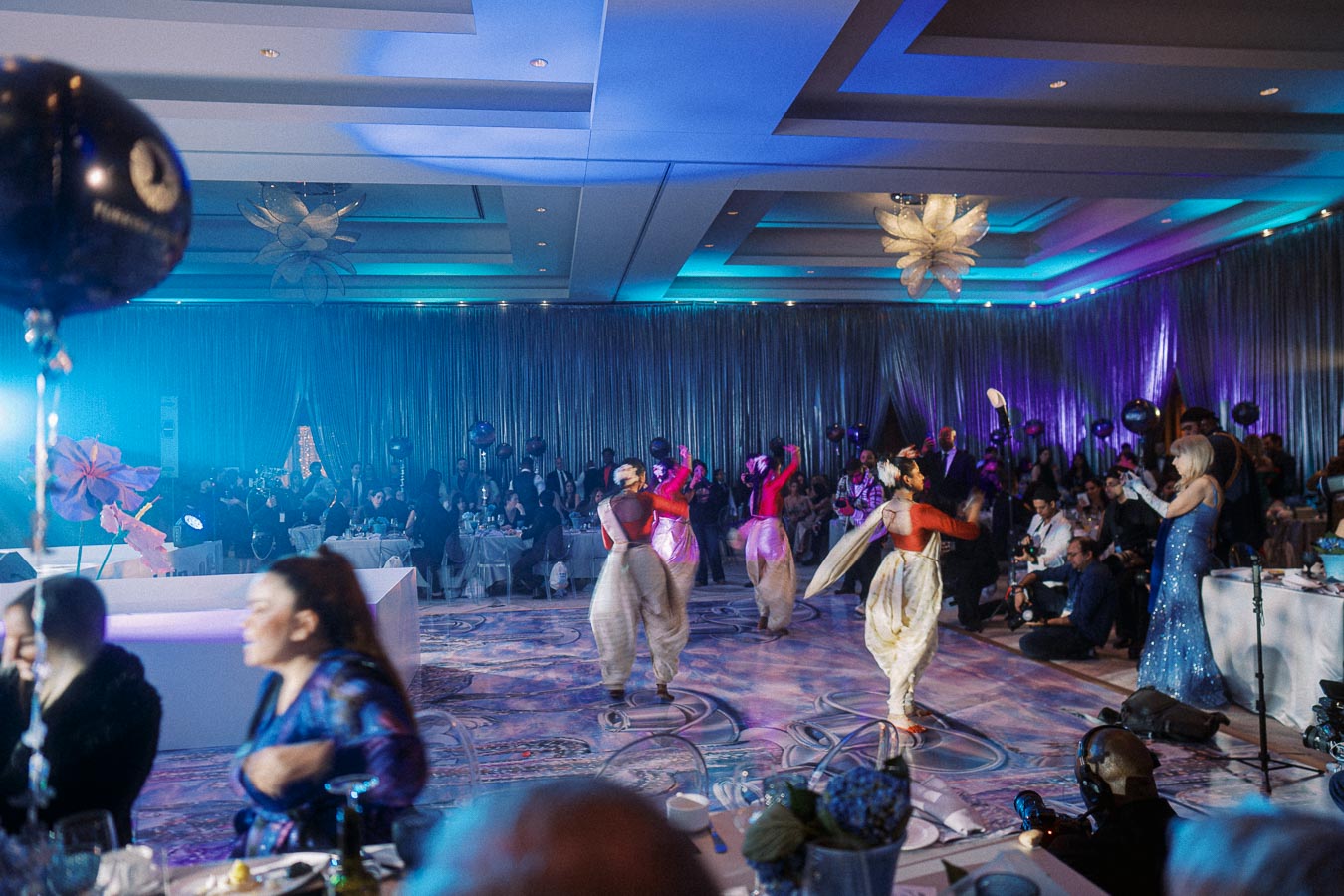 A vibrant and lively indoor event featuring traditional dancers in colorful costumes performing on a beautifully lit stage, surrounded by an audience seated at elegantly decorated tables with draped curtains and ceiling decor enhancing the festive atmosphere.
