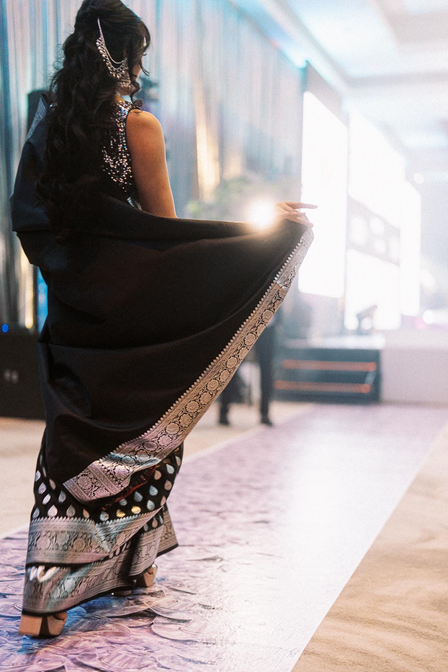 Elegant woman in a black saree with intricate silver embroidery walking gracefully down a runway with a blurred background.