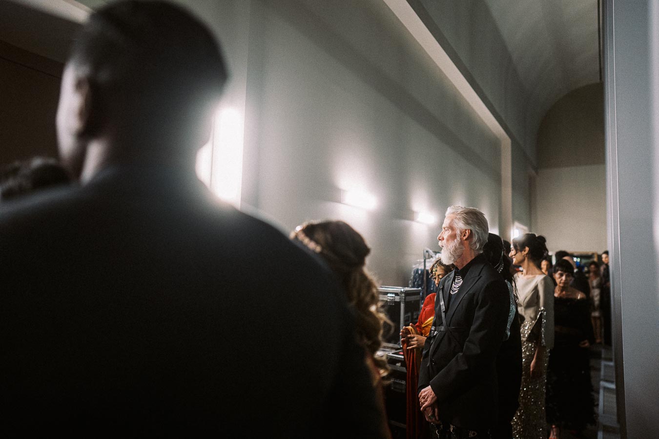 Backstage at a fashion show, a diverse group of elegantly dressed individuals await their turn under bright lights. The atmosphere is focused and anticipatory, capturing the behind-the-scenes preparation before hitting the runway.