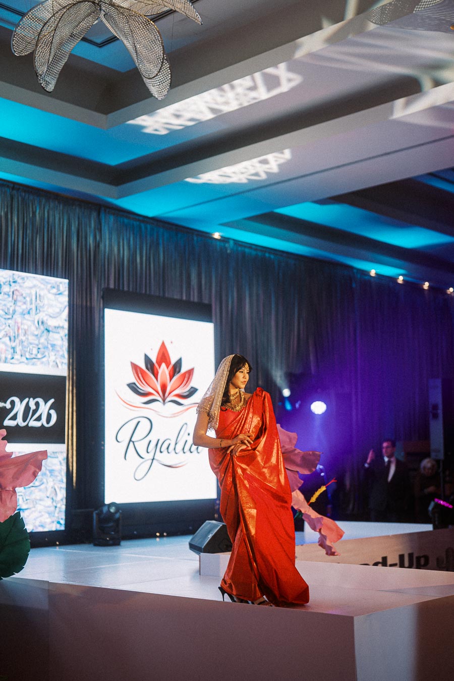 A model in a vibrant red saree walks the runway during a fashion show, with a backdrop displaying the Ryalix brand logo and decor.
