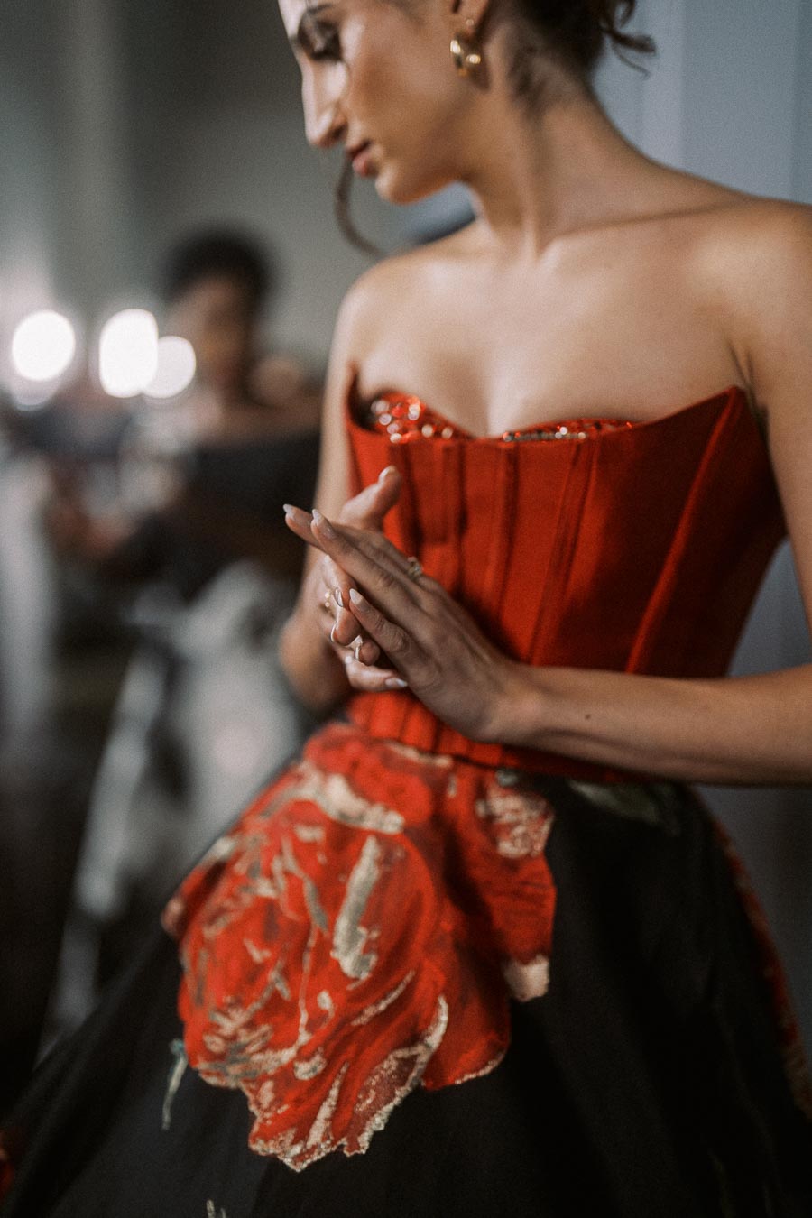 Elegant woman in a red corset gown with floral patterns, focusing on her clasped hands in a softly lit room.
