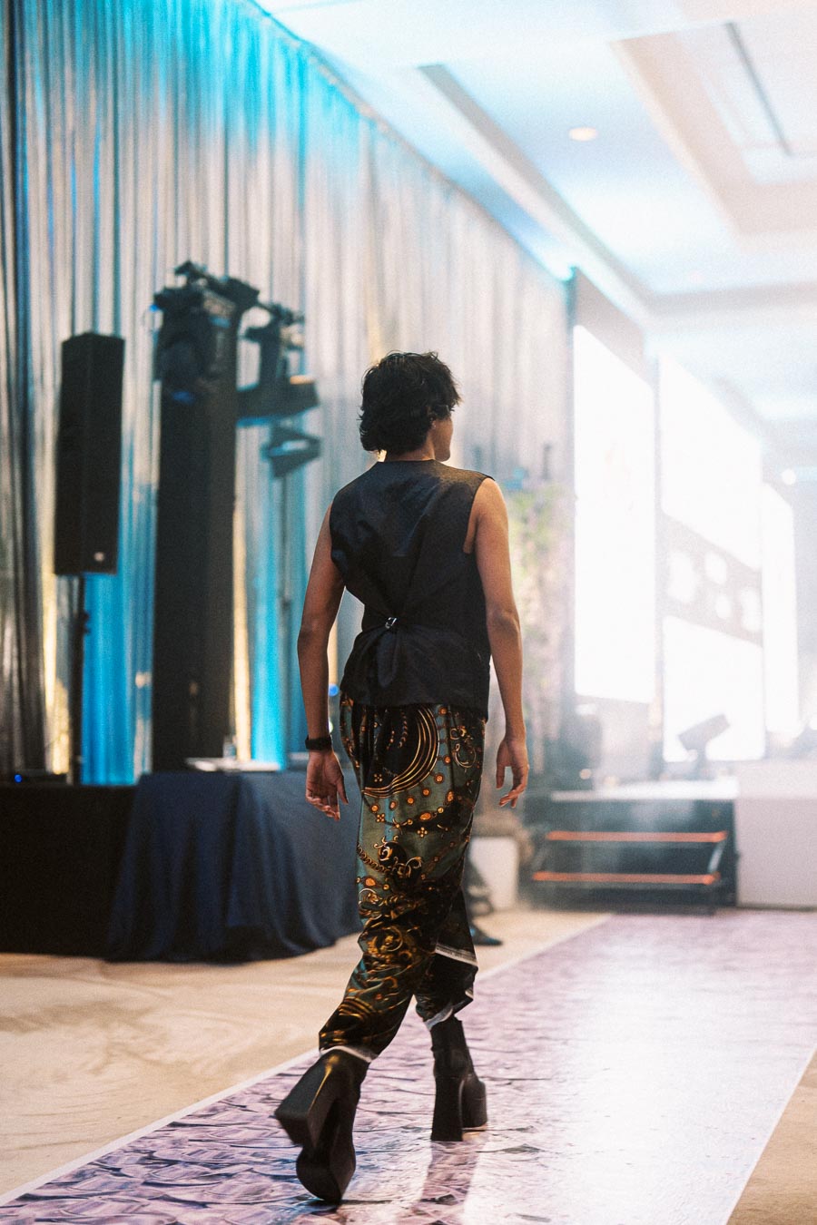 Fashion model walking down a runway in a stylish outfit featuring intricate patterns, high boots, and a sleeveless top at an indoor fashion show event.