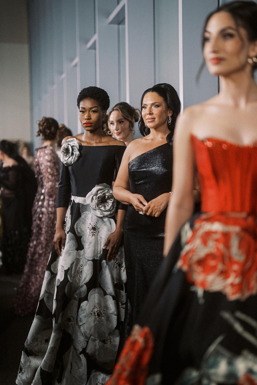 Elegant fashion models showcasing designer evening gowns at a glamorous indoor event, highlighting modern couture styles with floral and sequined details.