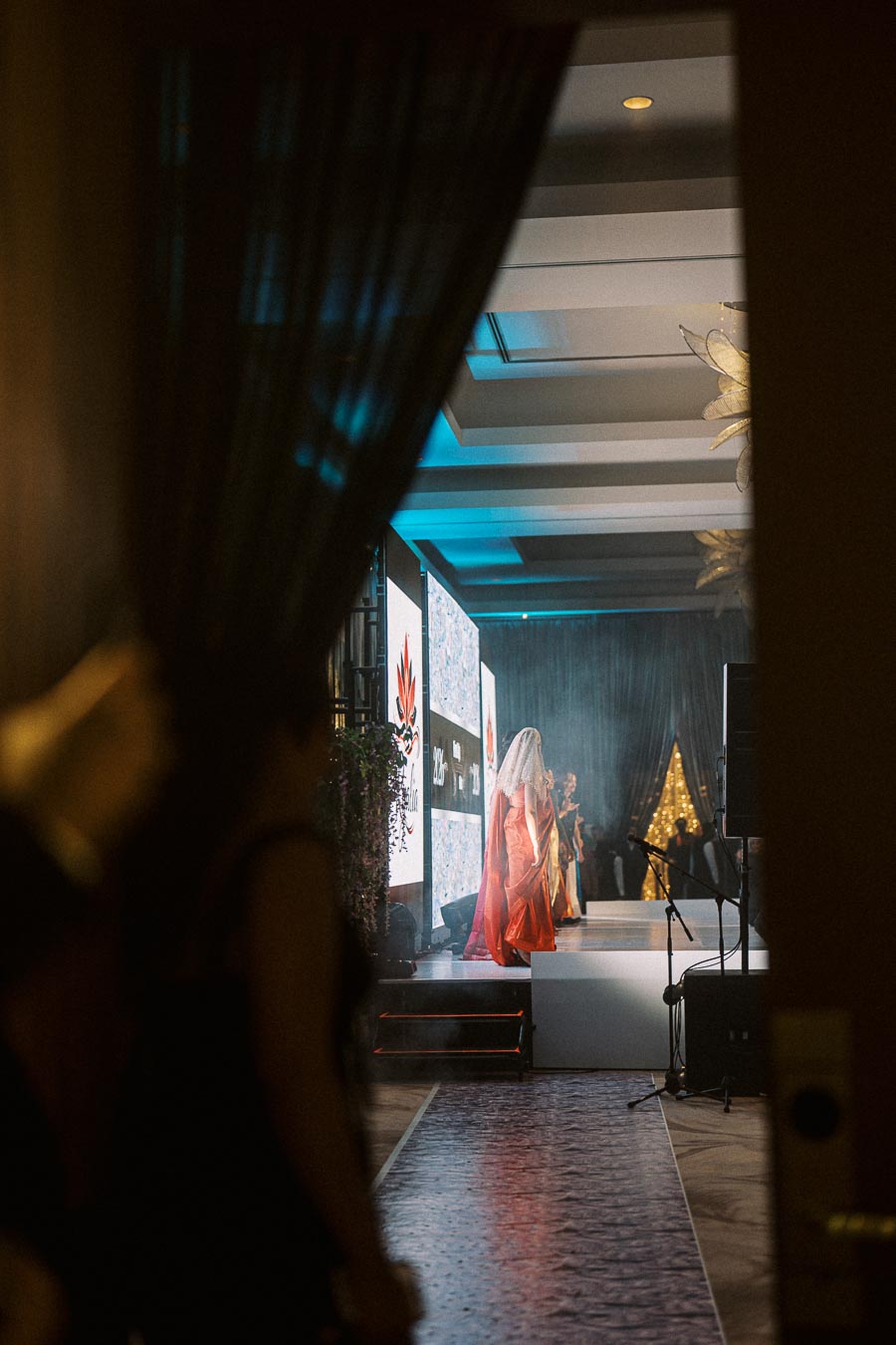 A glamorous fashion show with models wearing elegant attire walking down the runway, viewed through a doorway with dimly lit surroundings, highlighting the event's exclusivity and sophistication.
