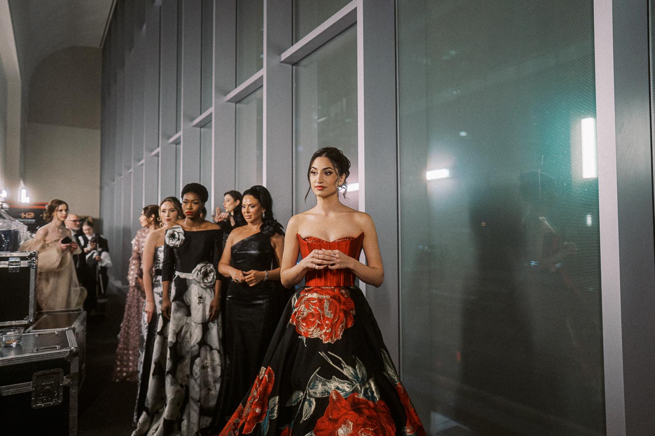 A group of elegantly dressed women stand backstage at a fashion event, featuring diverse evening gowns in a range of styles and colors, highlighted by a woman at the forefront wearing a striking red and black floral dress.