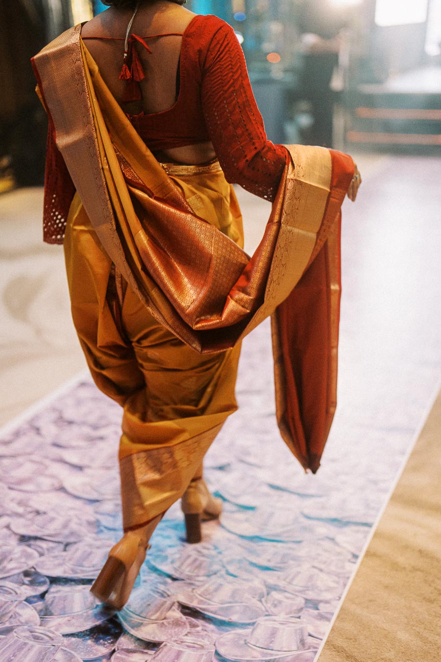 Woman in traditional Indian saree with intricate draping walks gracefully on a runway, showcasing elegant fashion and cultural attire.