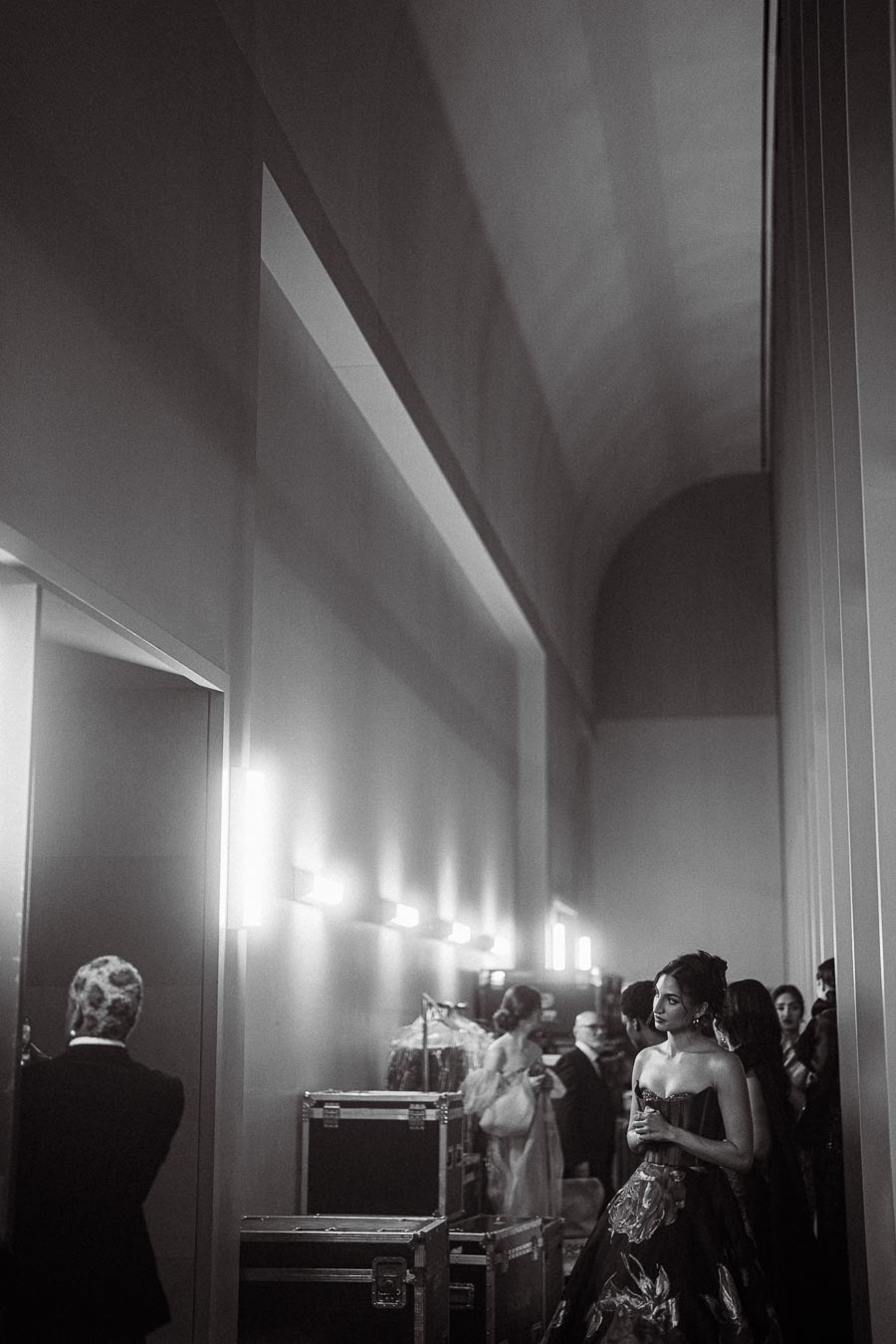 Black and white backstage scene with elegantly dressed individuals preparing for a fashion show, featuring women in stunning gowns and men in formal attire, surrounded by equipment cases in a well-lit corridor.