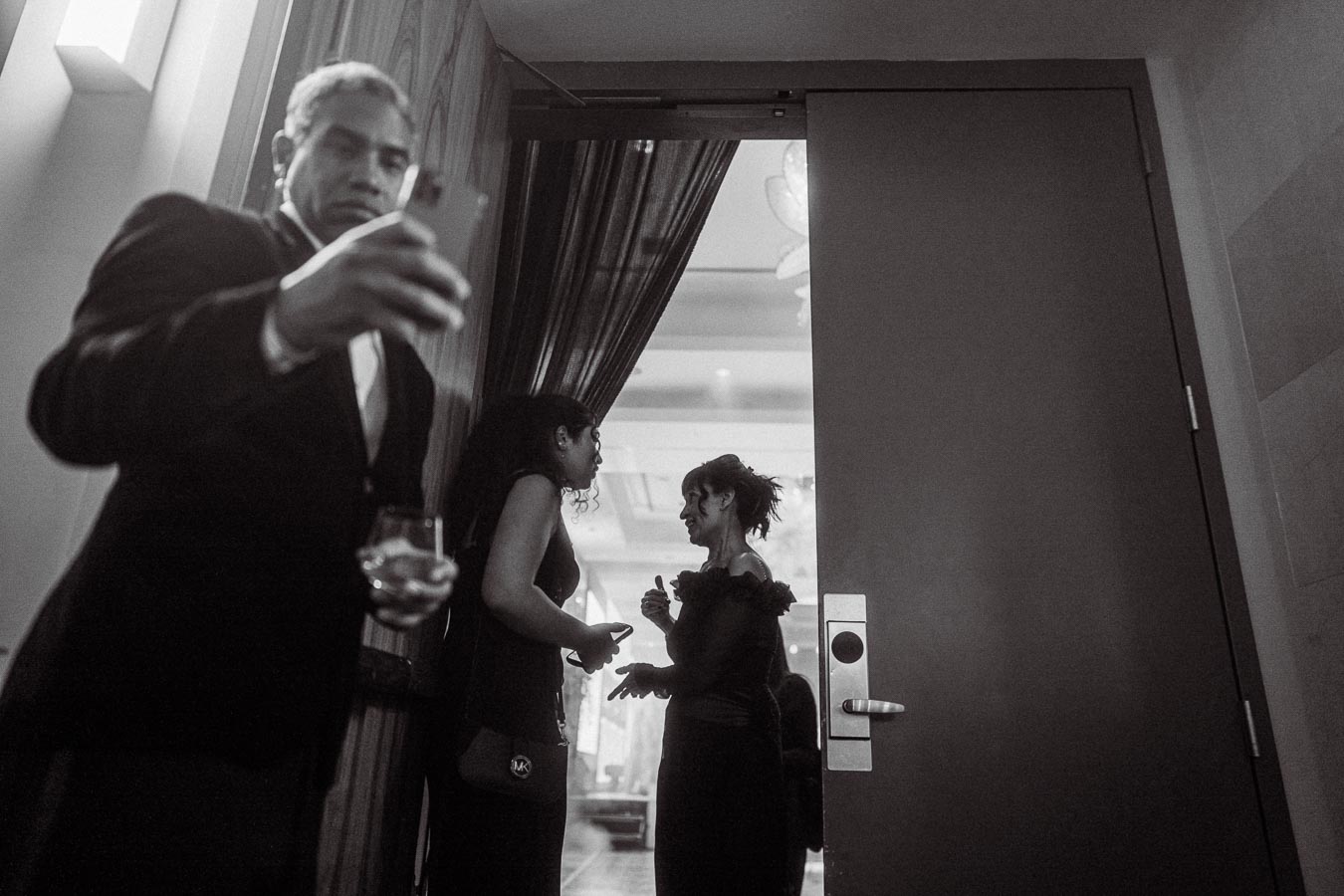 Black and white photo of a man in a suit taking a selfie with a smartphone, while holding a drink, near two women engaged in conversation at an elegant indoor event.