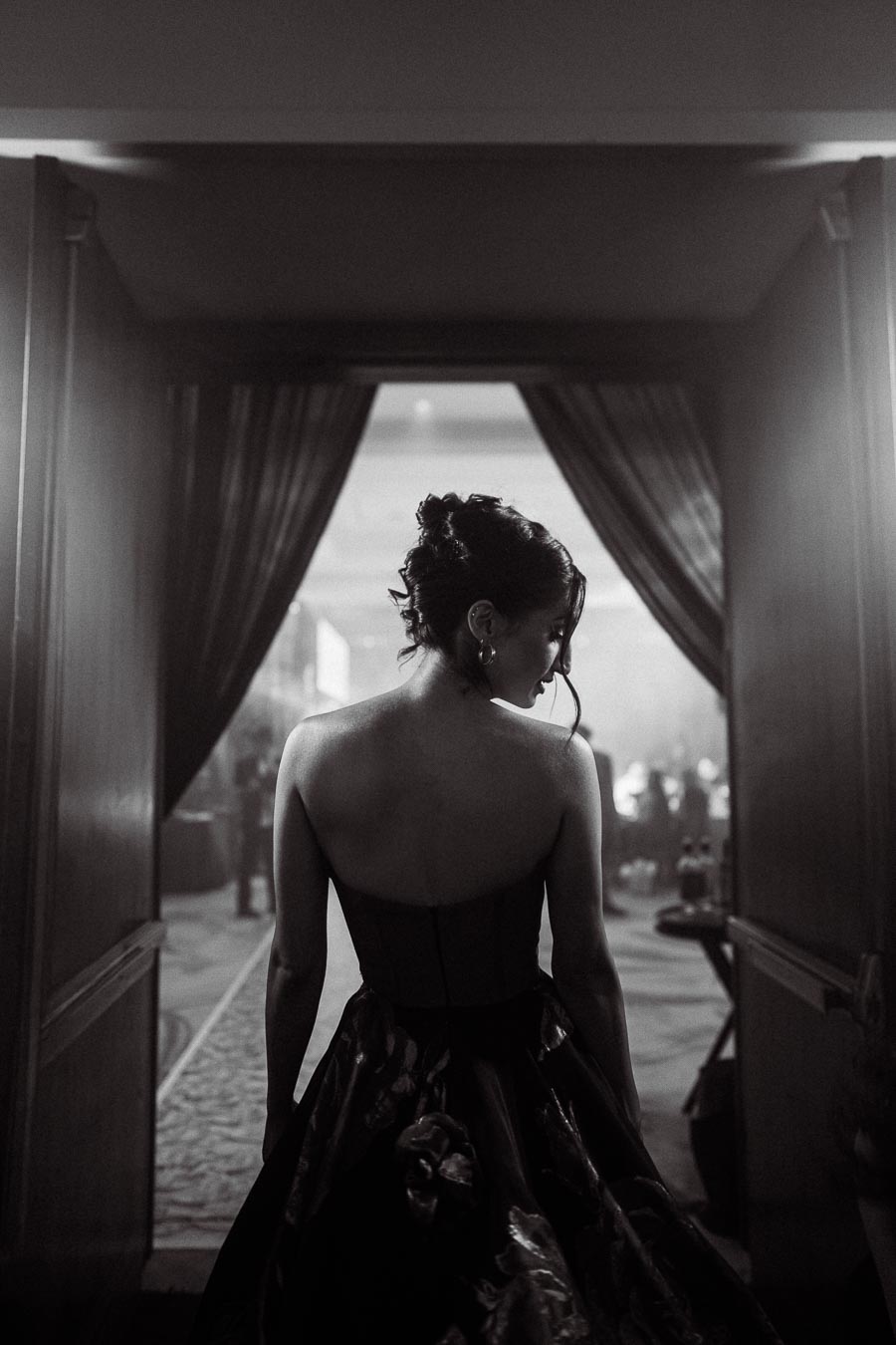 A woman in an elegant evening gown with floral details stands with her back to the camera in a dimly lit hallway, framed by curtains.