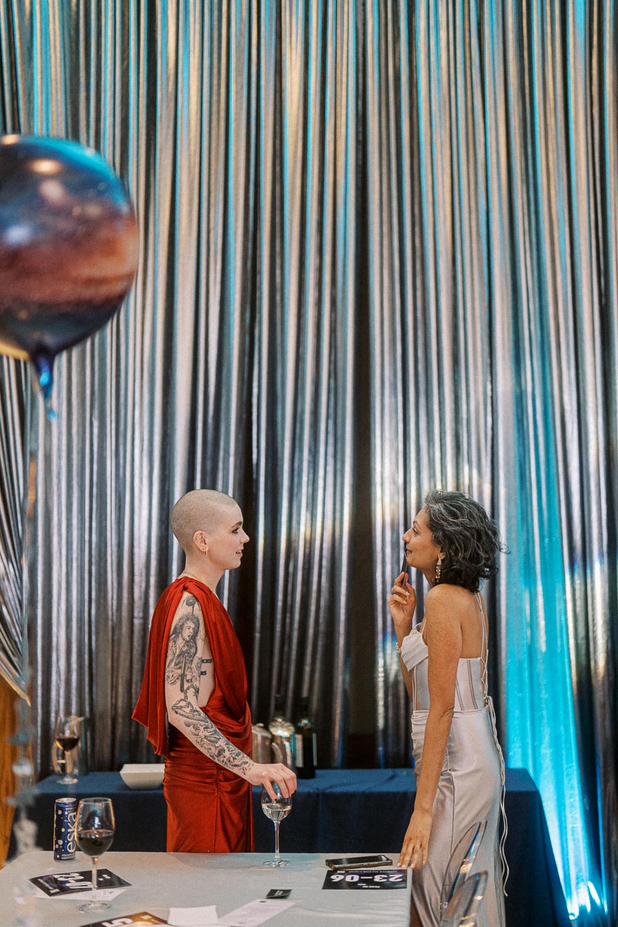 Two elegantly dressed individuals engage in conversation at a sophisticated event, set against a backdrop of shimmering drapes. A table with wine glasses and decorative elements is in the foreground, creating a chic and festive atmosphere.