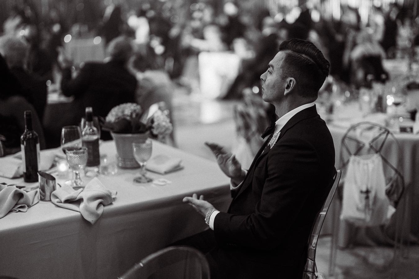 Black and white photo of a man in formal attire sitting at an elegantly decorated table during a social event, with wine glasses and a floral centerpiece visible, surrounded by other guests in a blurred background.