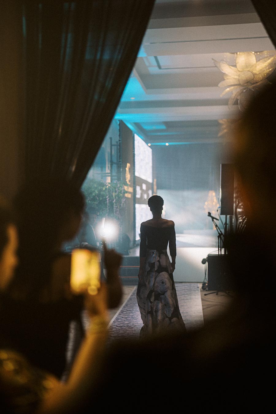 Fashion model walking on a dimly lit runway during a glamorous event, surrounded by onlookers capturing the moment with phones, under a dramatic ceiling light.
