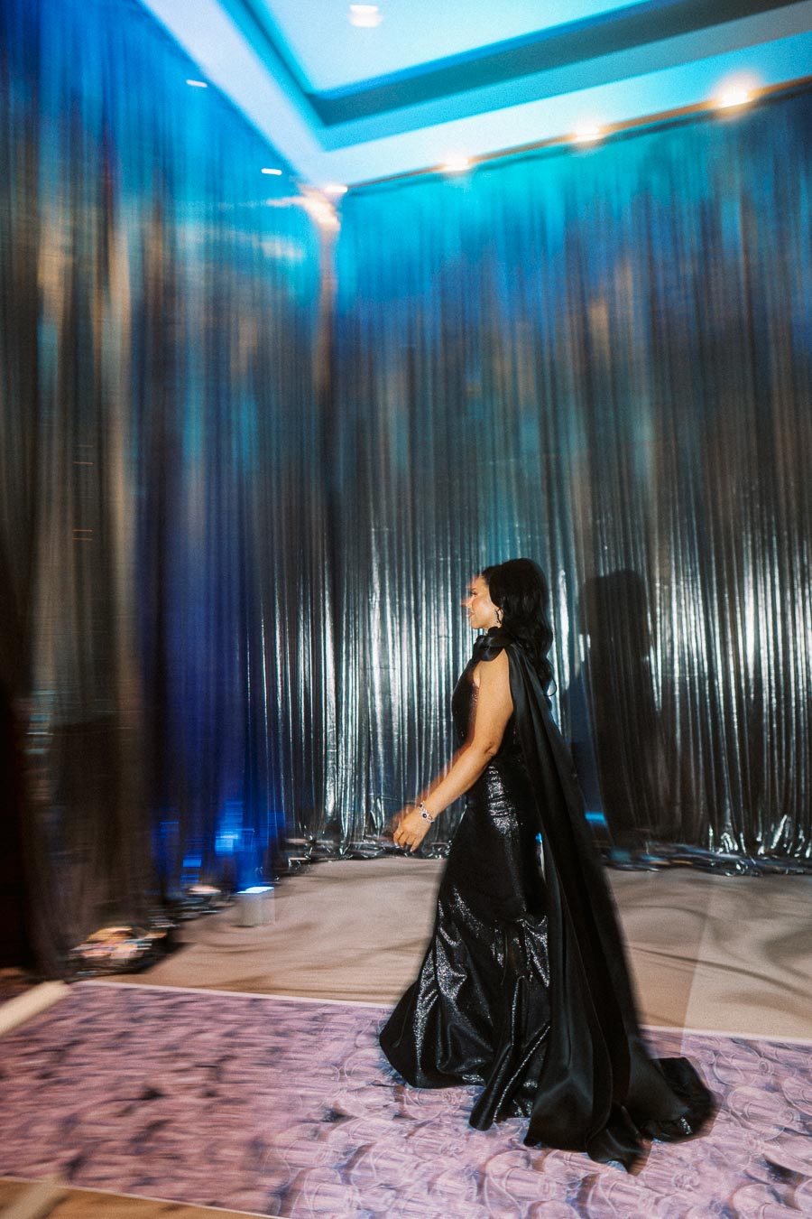 Elegant woman in a glamorous black evening gown walking by shimmering metallic drapes, under a modern ceiling with blue lighting.